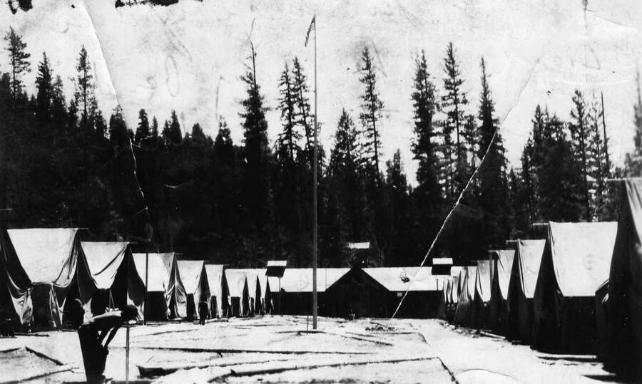 A view of Camp Four Corners, F-164, Company 533, Priest River, Idaho. Writing on the photo reads: 'CCC Camp 533 F-164 Priest River, Idaho.' The writing on the back of the photo reads: 'a picture of our street. The guy bending over is getting a drink. The building on the end is where we eat.'