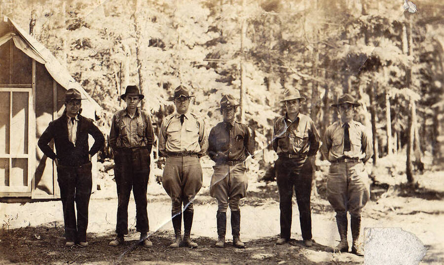 Forest and military personnel from camp F-164, Company 533, Priest River, Idaho. Writing on the photo reads: 'officers and forestry men'. Writing on the back of the photograph reads: 'Left to R. [Stnd?] J. McCormick, Oscour Webkiny (Camp Superintendent), [Stnd?] Westfall, Captain Meldrum, a forest ranger (don't know his name) Chaplain McDonnell.'