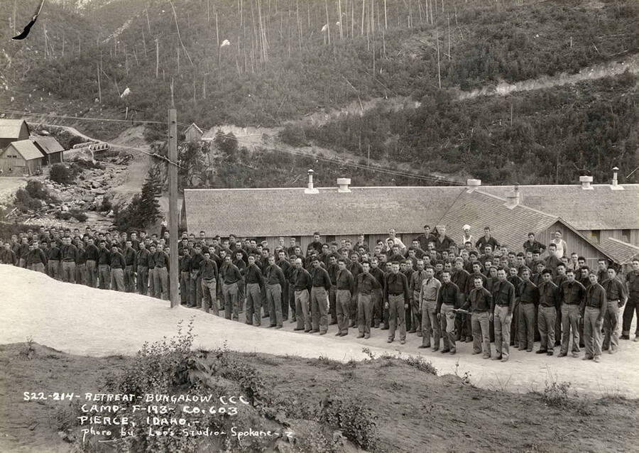 Group photo of uniformed CCC men standing at attention during retreat of the flag at Bungalow CCC Camp, F-193. Writing on the photo reads: 'Retreat Bungalow CCC Camp F-193 Company 603 Pierce, Idaho photo by Leo's Studio, Spokane.' Back of photo reads: 'Pierce, Idaho Bungalow'.