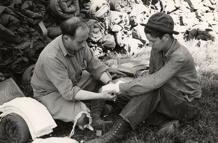 Photo of a CCC man getting his hand bandaged by another man in front of a wall of sleeping bags at the CCC Spirit Lake Fire Camp. Between the two men is a cigarette box that reads: 'Lucky Strike A Light Smoke'.
