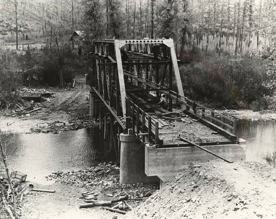 View of the McPherson bridge under construction over the Coeur d'Alene River in the Coeur d'Alene National Forest, McPherson Ranch can be seen in the background. Back of photo reads: 'McPherson Bridge over Trail Creek largely erected by CCC boys. McPherson Ranch in background. 1934'. Stamp on back of photo reads: 'Museum of North Idaho P.O. Box 812 Coeur d'Alene, Idaho 83814'.