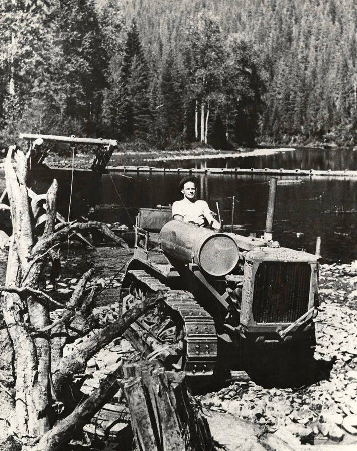 CCC man operating a cat tractor pulling a drag line to dredge sand and rock from the bottom of the Coeur d'Alene River. Sand and rock needed to make concrete for the Sissons Bridge. Back of photo reads: 'Loading gravel, Sisson's Bridge Coeur d'Alene River 1933'. Stamp on back of photo reads: 'Museum of North Idaho P.O. Box 812 Coeur d'Alene, Idaho 83814'.