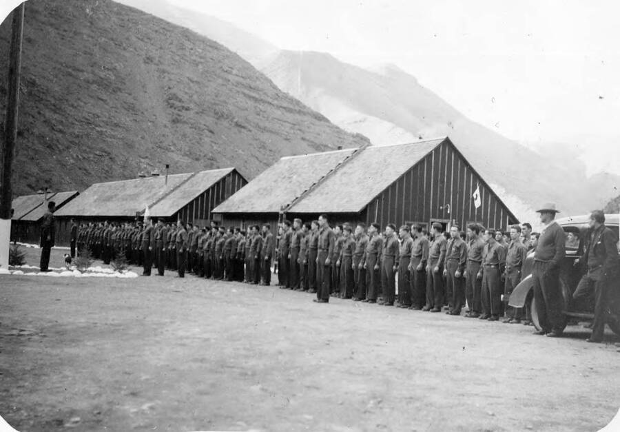 Group photo of uniformed CCC men standing at attention at retreat of the flag at CCC Camp Pollock, F-107, two miles from Riggins, Idaho. Back of the photo reads: 'Camp Pollock, 2 miles from Riggens. Retreat lowering of flag every night. C.J.P. St. Gertrude's Museum Civilian Conservation Corps'.