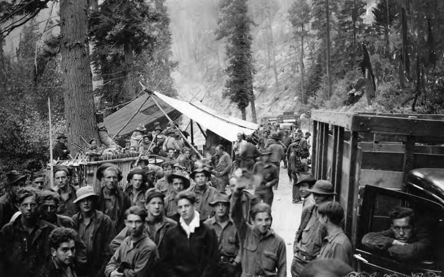 Group photo of a large group of CCC men at a fire camp at the mouth of Droogs Creek, on the South Fork of the Clearwater River below Golden, Idaho. Photo taken on August 10, 1936. Back of photo reads: 'This is the same place as other view. Early morning of Aug. 10 1936. Tools are arranged in racks, also a tent [_ ] is spread over cook stove. As near as I can remember and also from reference. Isa Duncan is in charge of the tool rack. At mouth of Droogs Creek, on South Fork of Clearwater. Note telephone on tree in foreground. Fire Camp at mouth of Droog Creek South Fork of the Clearwater. Below Golden Credit given to A.C. Duncan St. Gertrude's Museum Civilian Conservation Corps'.