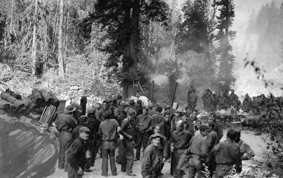 Group photo of a large group of CCC men at a fire camp at the mouth of Droogs Creek, 12 miles upriver from Castle Creek Station, four miles downriver from Golden, Idaho on the South Fork of the Clearwater River. Back of the photo reads: 'Fire Camp at mouth of Droogs Creek on South Fork. approx. 12 miles upriver from Castle Creek Station and 4 miles downriver from Golden Just around the bend of road beyond the last men is the large over-hanging reach. The year is 1936 as near as I can remember and believe is correct. Both Camp O'Hara CCC boys and Red River CCC Camp were on this fire. Camp Red River had winter quarters at Camp 107 Riggens, Idaho. Photo taken by C. J. Poxleitner and [_] by A.C. Duncan. Picture shows crews [rest] after breakfast [with]. St. Gertrude's Museum Civilian Conservation Corps.'