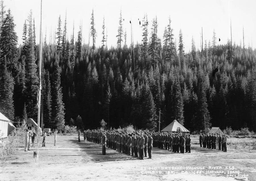 CCC Company 1655 standing at attention in front of an officer and the flagpole. The flag is being raised. The writing on the photo reads: 22-060 Retreat - Palouse River CCC. Camp F-185 - Co-1655 - Harvard, Idaho Leo's Studio' The writing on the back of the photo reads: '25-13-50 1930's [unidentified name] of N. Idaho CCC Camp Harvard Palouse R.'
