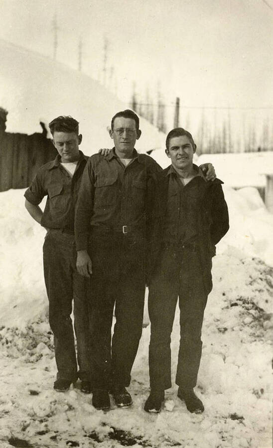 Three CCC men in Camp S-260 Big Creek  CCC Camp, Company 244. There are barracks in the background and snow on the ground.