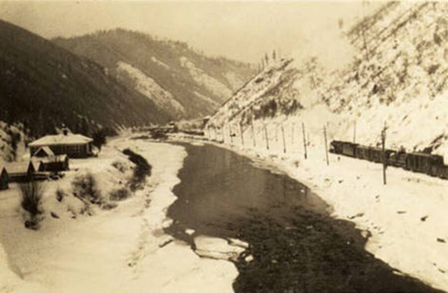 A partly frozen-over river runs between a CCC camp and a railroad track at the bottom of a valley. The back of the photo reads: ''Avery Valley' Feb. 1939'.