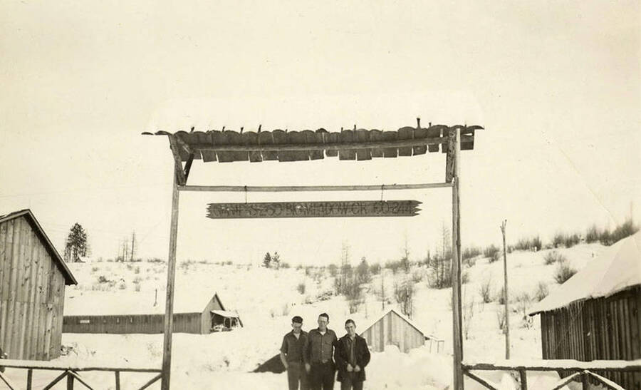 Three CCC men stand underneath a sign for Camp S-260 Big Creek  CCC Camp, Company 244. There are barracks in the background and snow on the ground.