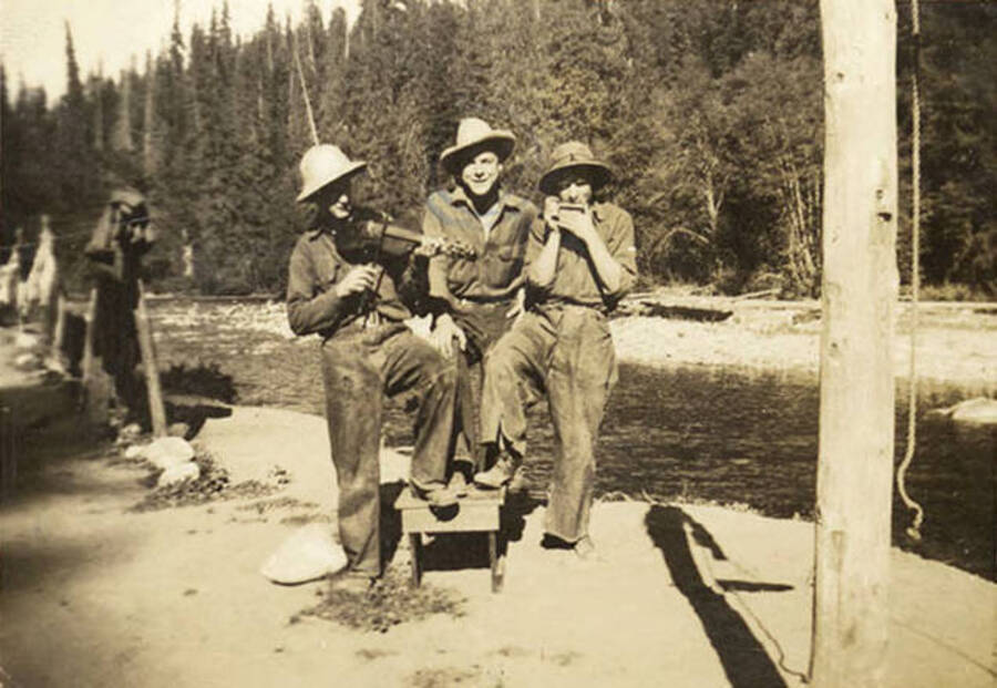 Three CCC men standing in front of a river at their CCC Camp. Two of the men are playing instruments. Back of the photo reads: 'Doug Eier Canter, Bowles Cabin - Tents put up along the Little North Fork River'.