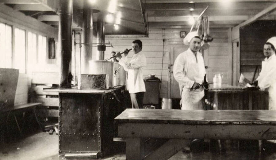 Four CCC men in the main kitchen of CCC Camp F-187. Two are sharpening knives and one is stirring the contents of a large pot. Back of photo reads: 'Kitchen - main camp August 29, 1938.'