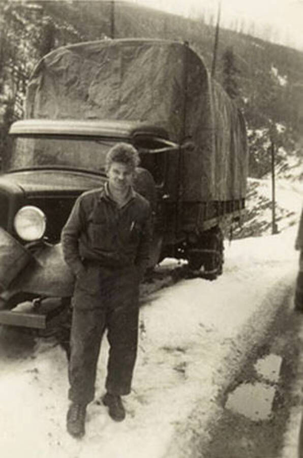 A CCC man stands in front of a covered truck parked on the side of the snow covered road.