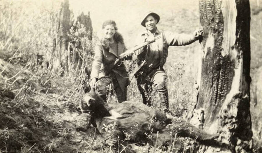A Forest ranger and his wife pose with a bear carcass. The ranger is leaning against a fire scarred tree. Back of the photo reads: 'A Ranger and his wife and their catch. Idaho 1938.'