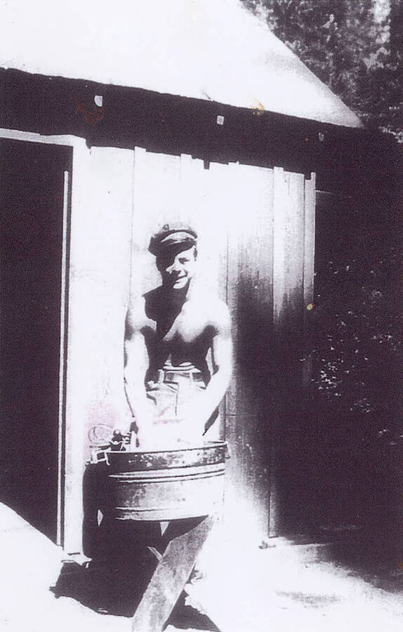 A shirtless CCC man standing in front of a wooden structure with his hands in a bucket that is atop a stool. Likely washing clothes.