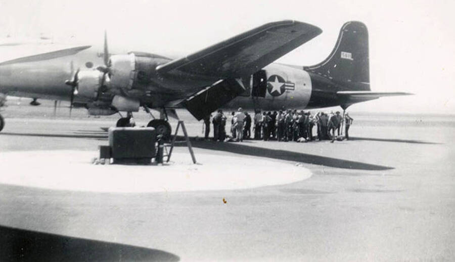 Several CCC men standing outside of an airplane. The hatch behind the wings is open and there is a ladder leading up to it. The back of the photo reads: 'firefighters loading Lewiston '49'.