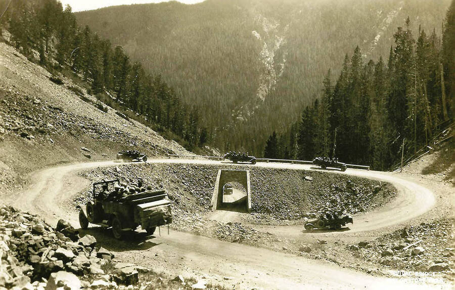 Six trucks can be seen driving on a road that circles back underneath itself. Forest covered hills dominate the background. The back of the photo reads: 'Yellowstone Park 1920's from Dr. Bowman'.