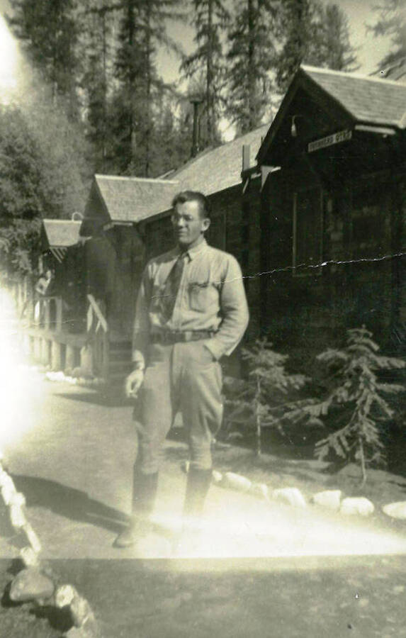 A Forest Ranger stands in front of CCC buildings at Camp Devil's Elbow, F-154. Back of photo reads: 'Forest Ranger at CCC Camp 'Devils Elbow' 6 or 7 miles above us on North Fork of the Coeur d'Alene River Pritchard, Idaho'.