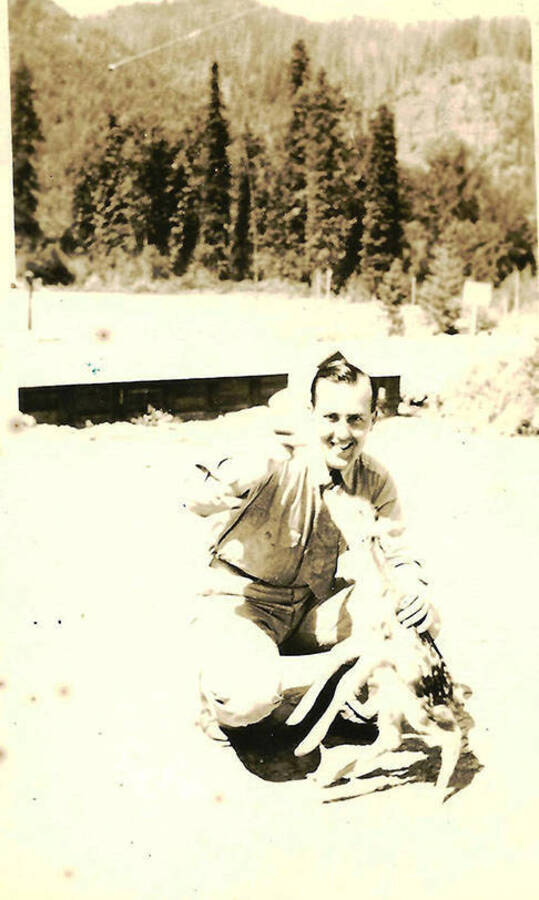 A CCC enrollee poses feeding a fawn from a bottle. CCC Camp Big Creek #2, F-132. The back of the photo reads: 'Feeding our pet deer (fawn)'.
