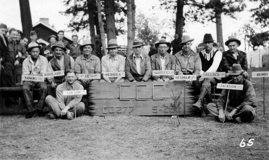 Group photograph of the Local Experienced Men (LEMS). The signs in front of each man read: 'Towns, Rowland, Ahlson (seated), Ax, Heikkila, (unidentified man), Morefield, Redman, Allred, Jackson (seated), and Henry. The large sign reads: 'C.C.C. Company 277 S-223.' Courtesy of John Boydstun.
