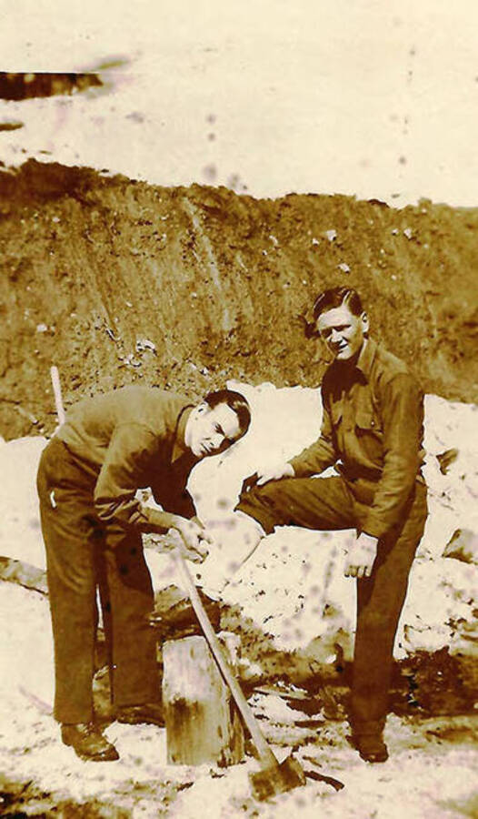 Two CCC enrollees posing for a photo. One is bandaging the other's leg, which is propped up on a stump. There is also an ax leaning against the same stump. CCC Camp Big Creek #2, F-132. Back of photo reads: ''cutting up' a little wood for barracks stove. Not supposed to cut your leg.'