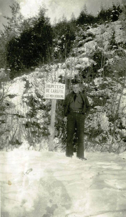 A CCC man stands in the snow in front of a sign that reads: 'Hunters Be Careful CCC Men Working'. CCC Camp Big Creek #2, F-132. Back of the photo reads 'George Petrisko from Steubenville CCC Co 531 Pritchard, Idaho 1939'.