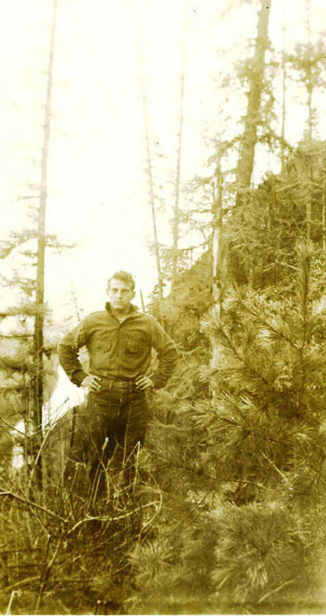 A CCC man stands in a stand of trees in the woods.CCC Camp Big Creek #2, F-132. Back of the photo reads: 'Hillsides are a little steep in this part of Idaho.'