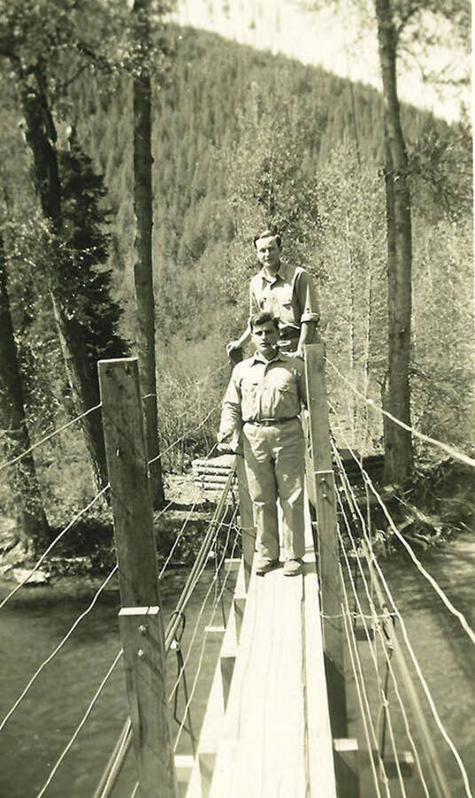 Two CCC men standing on bridge above a river. CCC Camp Big Creek #2, F-132. The bridge also holds a water line that feeds the camp. Back of the photo reads: 'water line over Uranus Creek near camp.'
