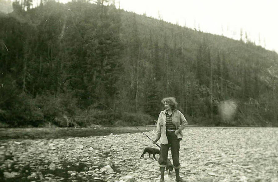 A woman on the banks of a river with two dogs and a fishing pole in her hand. CCC Camp Big Creek #2, F-132. Back of the photo reads: 'She really caught some fish (trout)'