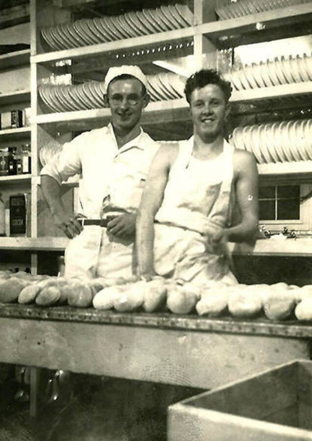 Two CCC men stand in the kitchen dressed in cooks outfits with rolls of dough on a table in front of them and lines of plates on shelves behind them. Back of the photo reads: 'Our bakers get the bread ready for a hungry mob'.