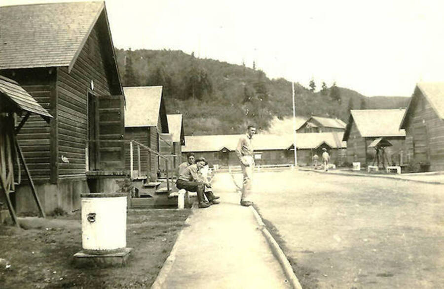 Three CCC men outside of the barracks with two others walking by in the background. The headquarters building can be seen in the background. CCC Camp Big Creek #2, F-132.