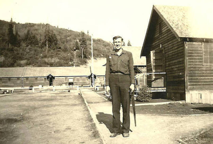 A CCC man, 1st Sergeant, poses in front of the barracks and headquarters building. CCC Camp Big Creek #2, F-132. Back of the photo reads: '1st Sgt. on company street at CCC Co 531'.