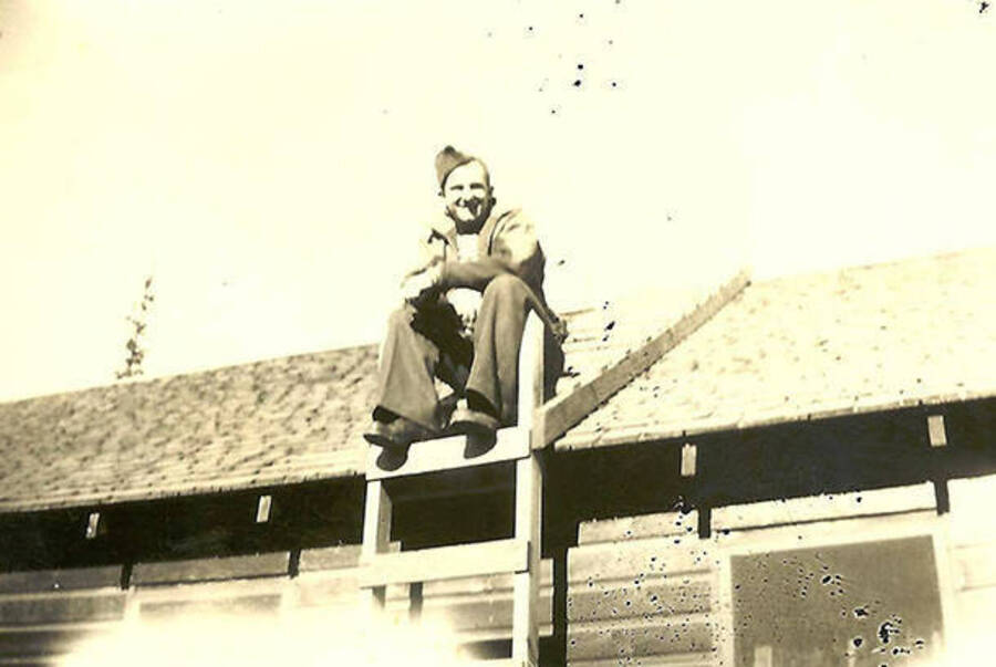A CCC man perches atop a barracks roof at CCC Camp Big Creek #2, F-132. Back of photo reads: 'High and dry. Checking the barracks roof'.