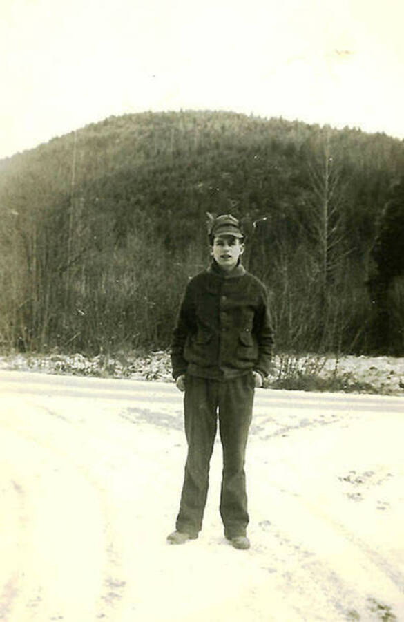 A CCC man poses on a snow-covered road, a tree-covered hill in the background. CCC Camp Big Creek #2, F-132. Back of the photo reads: 'Mike Misconish our driver of the team of Belgian horses we logged with. From the Steubenville area (mingo jet.)