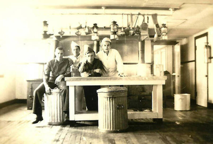 Four CCC men in the kitchen at CCC Camp Big Creek #2, F-132. Lanterns and kitchen utensils hang from the ceiling. One of the men perches on a trash can  while another can sits in front of the island counter.