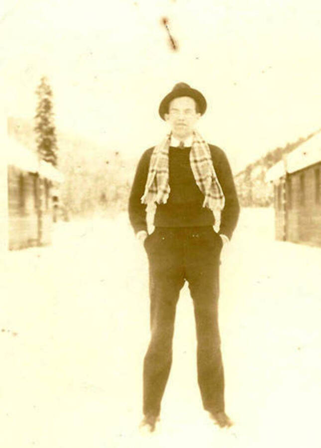 A man dressed in a shirt and tie, with a sweater over them, suit pants, scarf, and hat stands in-between the barracks. CCC Camp Big Creek #2, F-132.
