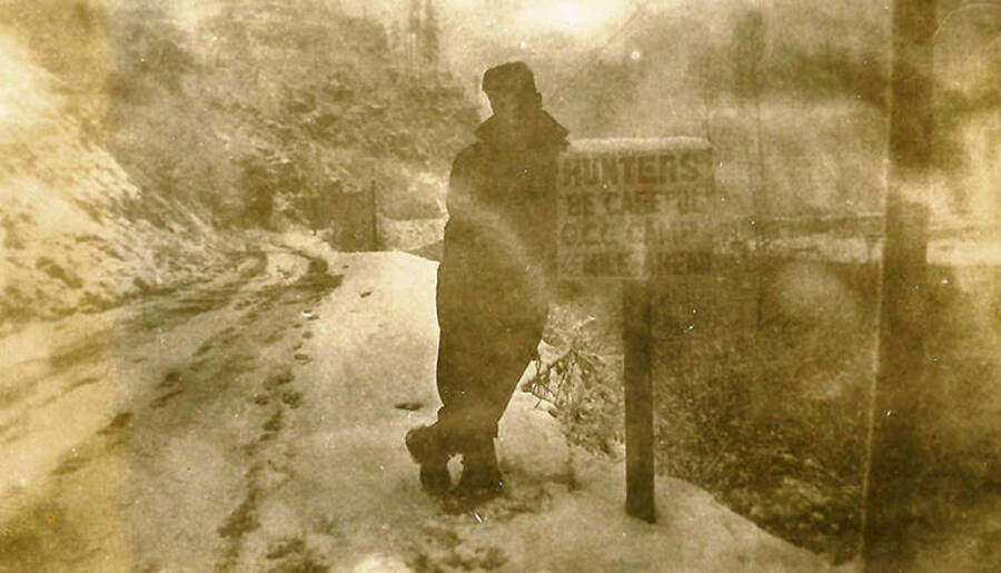 A CCC man stands in the snow next to the roadway by a sign that reads: 'Hunters Be Careful CCC Camp 1/2 mile ahead'. CCC Camp Big Creek #2, F-132. Back of the photo reads: 'Roads were not too great in winter. Taken just below camp at Big Creek.'