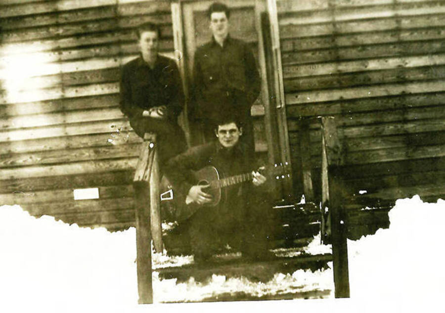 Three CCC men pose in front of the barracks in the snow with one of the men holding a guitar. CCC Camp Big Creek #2, F-132.