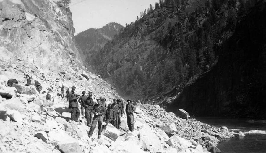 Men at work on a mountainside along a river. Potentially moving rocks to build a road.