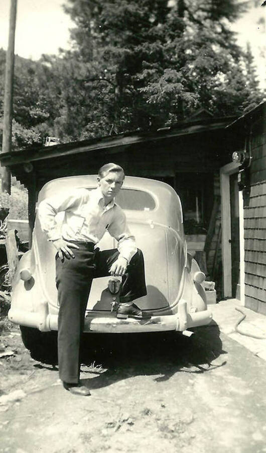 A CCC man poses with his foot on the bumper of an automobile at CCC Camp Big Creek #2, F-132.