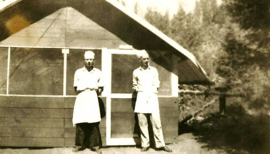 Two CCC cooks standing outside the mess hall at CCC Camp Big Creek #2, F-132. Back of photo reads: ''Cooks' outside our messhall'.