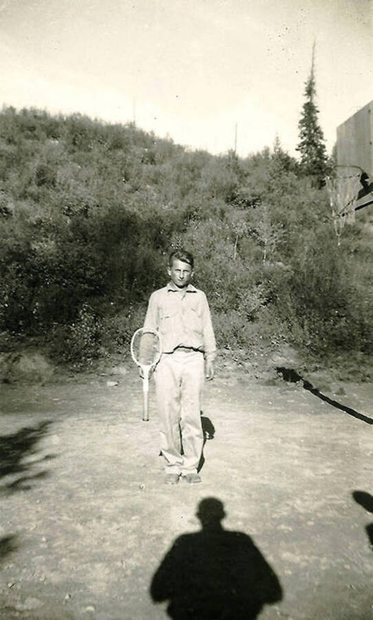 A CCC man poses with a tennis racket next to a basketball hoop. The photographer's shadow stretches across the foreground of the photograph. CCC Camp Big Creek #2, F-132.