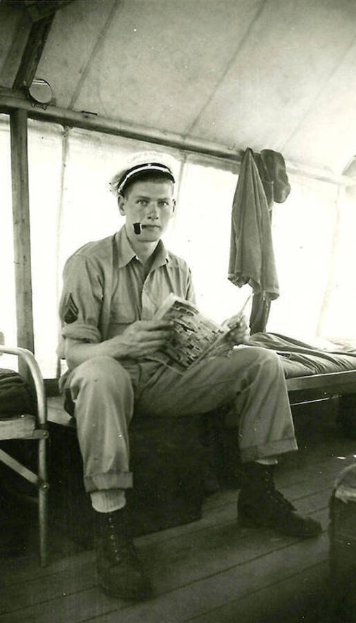 A CCC man sits on a chest in the barracks with a pipe and newspaper and/or comic. CCC Camp Big Creek #2, F-132.