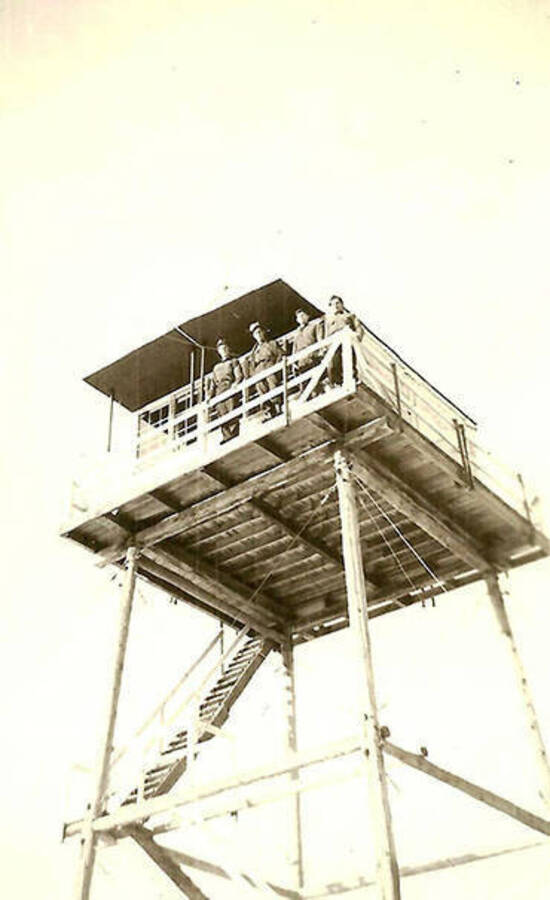 Four CCC men look down from a lookout tower near CCC Camp Big Creek #2, F-132. Back of the photo reads: 'Our fire tower on Uranus Peak.'