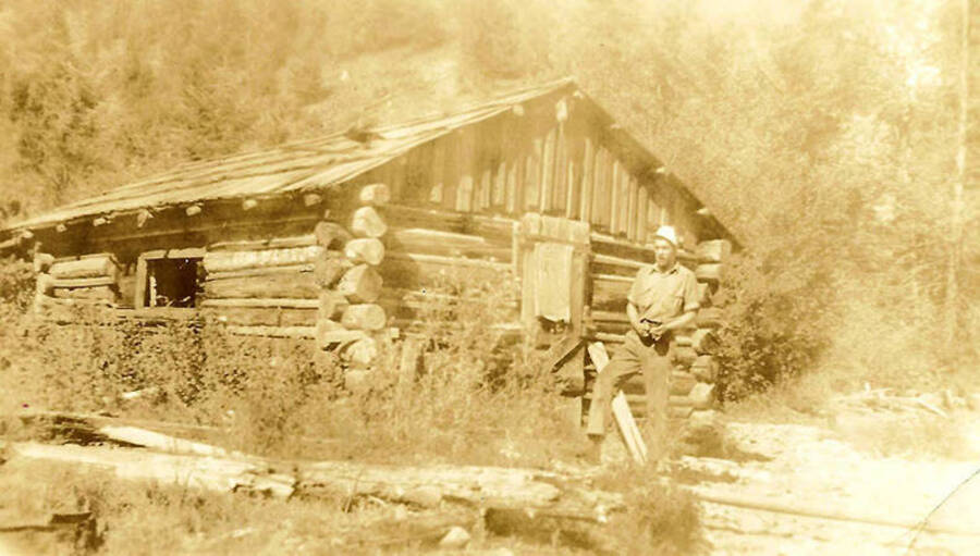 A CCC man poses in front of a log cabin.