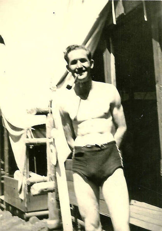 A CCC man in a swim suit smoking a pipe in front of  the tent barracks at CCC Camp Big Creek #2, F-132.