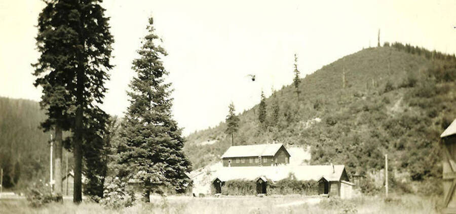 The Forest Service Office is shown against a hill in the middle of the forest. Back of the photo reads: 'View of the Forest Service office building with the infirmary in the background. Taken in 1933