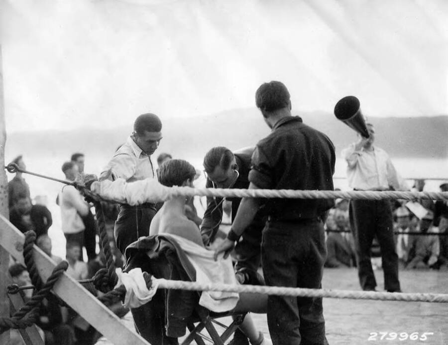 A boxing entrant in his corner, being medically examined while a man announces with a megaphone in the background at the CCC Carnival in McCall, Idaho. 'Boxing wound up the day's sports at the CCC Carnival in McCall, Idaho - 3 camps competing.'