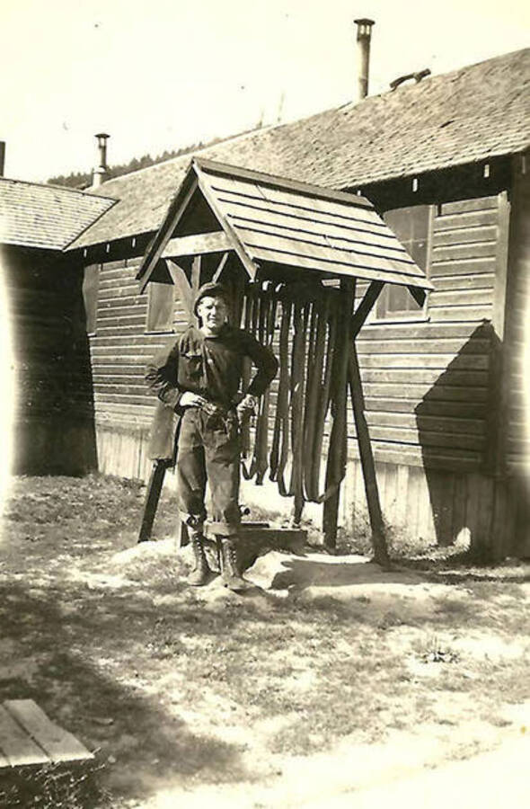 A CCC man poses for a photo next to the barracks at  CCC Camp Big Creek #2, F-132. He is standing in front of a small shelter with long loops hanging from a beam, presumably fire hoses. Back of the photo reads: 'Fire equip't at barracks'.