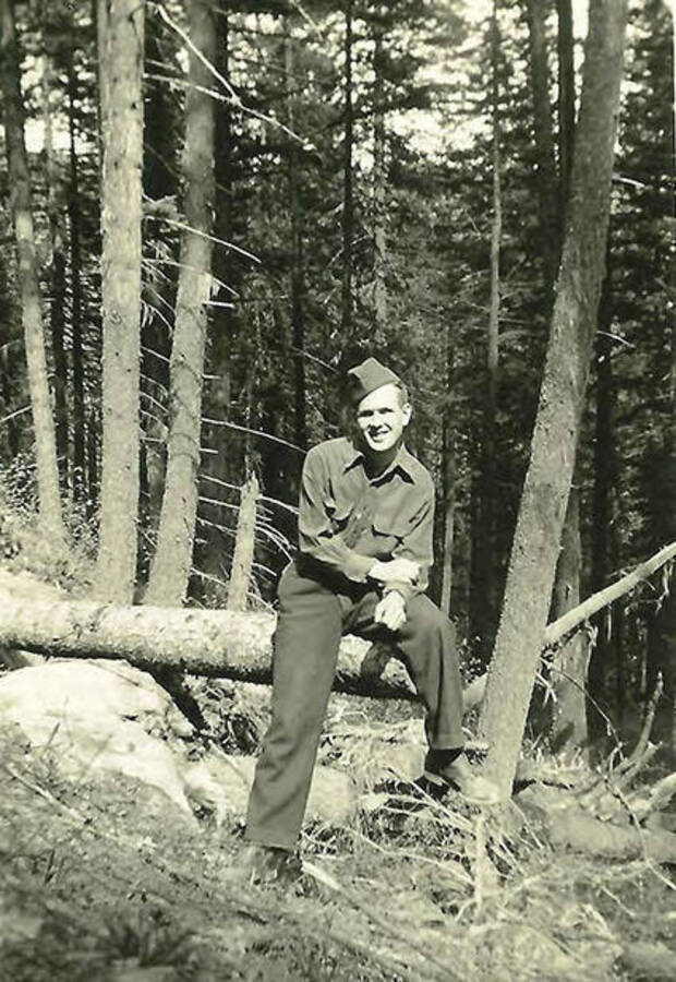 A CCC man sits on the edge of a log in the woods near CCC Camp Big Creek #2, F-132.