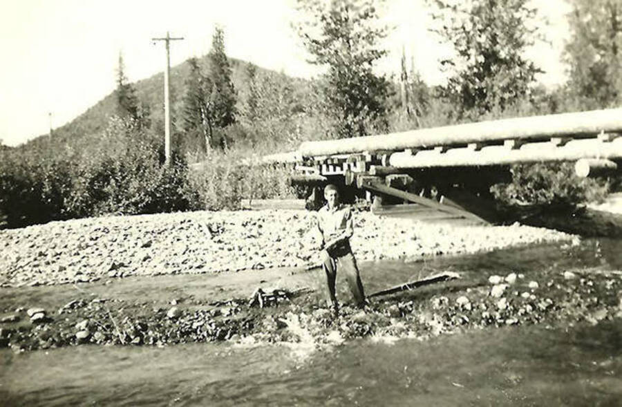 A CCC man stands in the middle of the creek next to a bridge near CCC Camp Big Creek #2, F-132. The back of the photo reads: 'Bridge over big Creek below camp'.